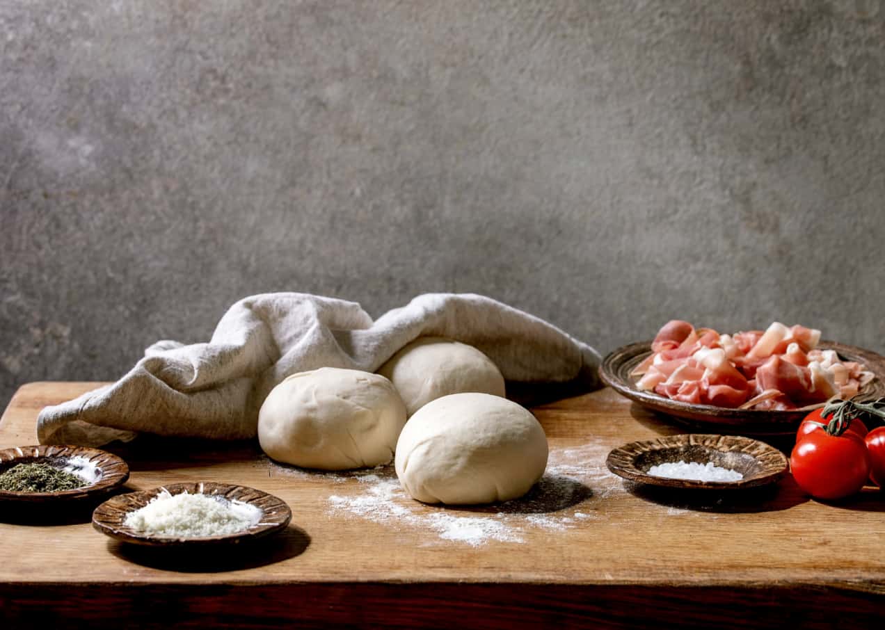 Foto di panetti di impasto su un tavolo di legno e con accanto dei piatti con sale, prosciutto, pomodorini